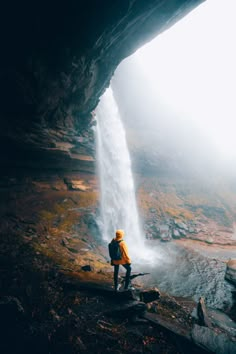 a person standing at the base of a waterfall looking out into the distance with fog in the air