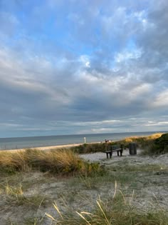 two benches sitting on top of a sandy beach next to the ocean under a cloudy sky