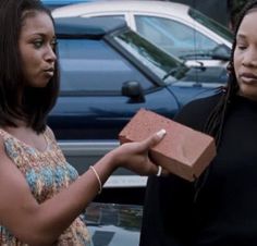 two women standing next to each other in front of a blue car and one is holding a brick