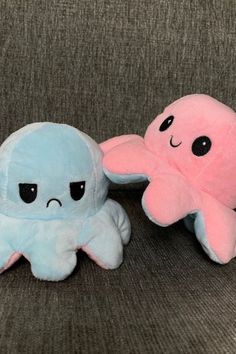 two stuffed animals sitting next to each other on a gray couch, one is pink and the other is blue