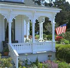 an american flag on the porch of a white house with flowers and shrubs around it