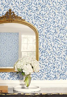 a vase with white flowers in front of a blue wallpapered mirror on a table