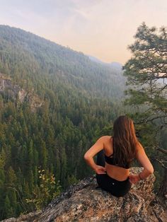 a woman sitting on top of a large rock next to a lush green forest covered hillside