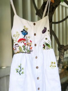 a white dress hanging from a tree with flowers and butterflies on it's chest