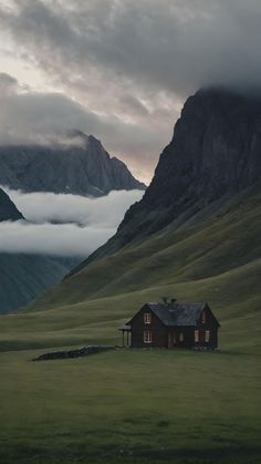 a house in the middle of a field with mountains in the background and clouds rolling over it