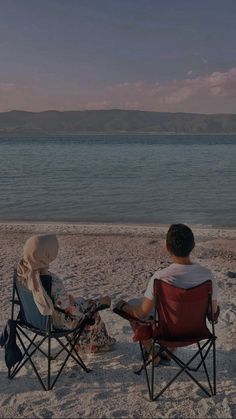 two people sitting in lawn chairs on the beach watching the sun set over the water