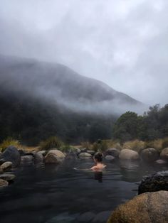 a man swimming in a hot tub surrounded by rocks and grass with mountains in the background