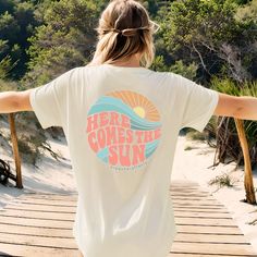 a woman walking across a wooden bridge with her arms spread out to the side while wearing a t - shirt that says here comes the sun