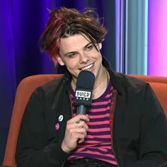 a person sitting in a chair with a microphone and smiling at the camera while wearing a striped shirt