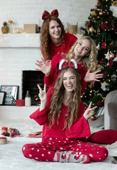 two women in matching christmas pajamas posing for the camera with their hands up while sitting next to a christmas tree