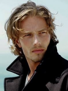 a man with long hair wearing a black jacket and looking off into the distance while standing in front of water