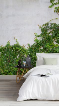 a bed with white sheets and pillows in front of a wall covered in green plants