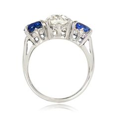 This antique three-stone engagement ring centers a 2.04 carat old European cut diamond, J color and VS1 clarity, set in prongs. The diamond is accented by two round cut sapphires with a combined weight of 2.56 carats, also set in prongs. The handcrafted platinum mounting features a fleur-de-lis motif on the under-gallery. This ring is from the Art Deco era, circa 1930.
The old European diamond measures approximately 7.83mm x 7.78mm x 5.19mm. The sapphires measure approximately 6.31mm x 6.16mm x Timeless Lab-created Sapphire Wedding Ring, Timeless Lab-created Sapphire Diamond Wedding Ring, Timeless Lab-created Sapphire Diamond Ring For Wedding, Classic Three Stone Jewelry With Lab-created Sapphire, Art Deco Three Stone Diamond Ring, Fine Jewelry Three Stone Lab-created Sapphire Ring, Three Stone Lab-created Sapphire Diamond Ring, Sapphire Three Stone Round Cut Diamond Ring, Three Stone Sapphire Diamond Ring With Round Cut