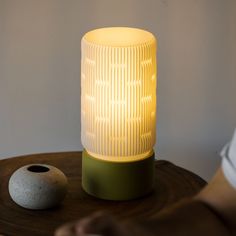 a white lamp sitting on top of a wooden table next to a gray vase and a person's hand