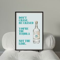 a bottle of alcohol sitting on top of a white pillow next to a framed poster that says don't chase be chased you're the tequila not the lime