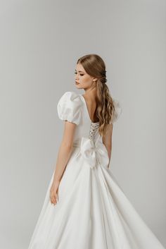 a woman in a white wedding dress with a large bow on her back, looking off to the side