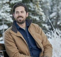 a man sitting on top of a wooden bench in front of some snow covered trees