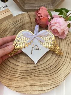 a hand holding a heart shaped brooch with angel wings on it and pink flowers in the background