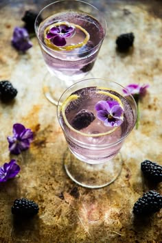 two glasses filled with liquid and blackberries on top of a wooden table next to purple flowers