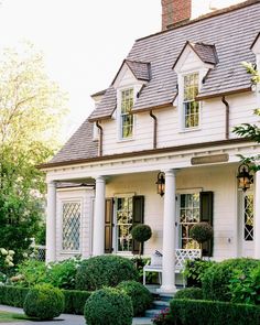 a white house with lots of plants and bushes around it's front porch area