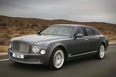 the front end of a grey bentley mulsanne driving on a road with mountains in the background