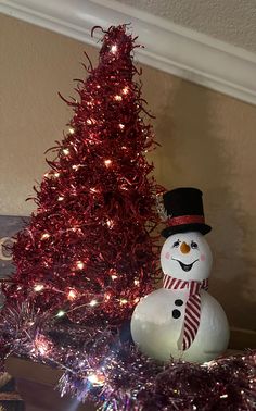 a snowman sitting next to a christmas tree with red tinsel and lights on it