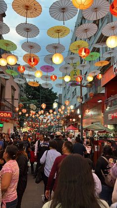 a crowd of people walking down a street next to tall buildings with umbrellas hanging from the ceiling