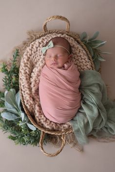 a newborn baby wrapped in a pink wrap laying on top of a basket with greenery