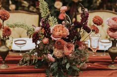 an arrangement of flowers in vases on top of a red table cloth with place settings