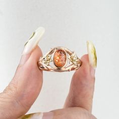 The Solstice Blossom ring draws its inspiration from the beauty of nature and the warmth of the sun. At its heart lies a radiant, oval-cut natural sunstone that shimmers with earthy brilliance. Flanking the centerpiece are two intricately carved flower motifs, each adorned with a sparkling round diamond at its center. The band is designed with a brushed, tree bark-like texture, evoking the rugged yet graceful essence of nature. This unique and artistic design is perfect for those seeking a custom engagement ring, promise ring, or anniversary ring that reflects natural beauty and timeless elegance. 𝗗𝗘𝗧𝗔𝗜𝗟𝗦𝗠𝗔𝗜𝗡 𝗦𝗧𝗢𝗡𝗘𝗚𝗲𝗺𝘀𝘁𝗼𝗻𝗲:Sunstone𝗖𝗼𝗹𝗼𝗿: Reddish brown𝗦𝗵𝗮𝗽𝗲: Oval Cut 6.0*8.0mm𝗖𝘂𝘁: Full𝗠𝗲𝗮𝘀𝘂𝗿𝗲𝗺𝗲𝗻𝘁: 1.5 ct approx𝗤𝘂𝗮𝗹𝗶𝘁𝘆: VS clarity grade? Earthy Engagement Rings, Gold Leaf Ring, Gold Leaf Rings, Rings Promise, Flower Rings, Leaf Engagement Ring, Rutilated Quartz Ring, Art Ring, Engagement Rings Bridal Sets