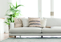 a white couch with pillows on it in front of a potted plant and window