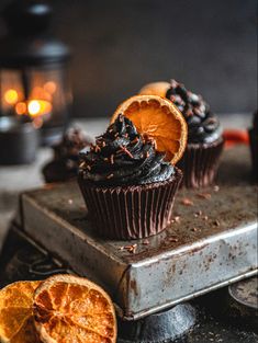 three chocolate cupcakes with orange slices on a metal tray next to some candles