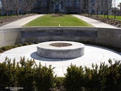 a circular concrete fire pit in front of a large building with trees and bushes around it