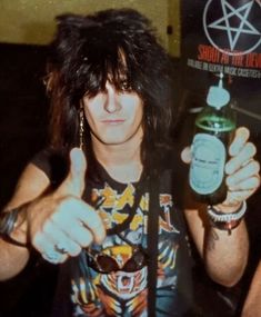 a man with long hair and piercings holding a bottle in his right hand while giving the thumbs up sign