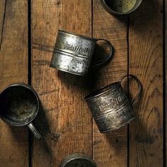 three old metal cups sitting on top of a wooden table next to two spoons