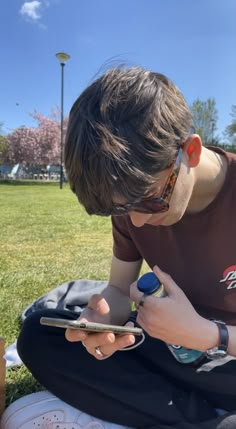 a young man sitting on the grass looking at his cell phone and writing something in front of him