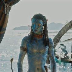 a man with blue paint on his body standing in front of the ocean and holding a stick