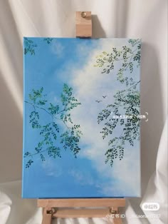 an easel with a painting on it holding a blue sky and green tree branches