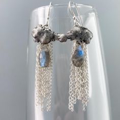 These handcrafted earrings feature dark clouds with an oxidized stippled texture above a downpour of bright silver chains in two different styles and teardrop briolettes of electric blue labradorite. They hang from handmade French hook earwires and measure 58mm/2.28" long by 17.5mm/0.69" wide.  To clean, wipe gently with a specially treated silver-polishing cloth (available in my Jewelry Care section or included with orders over $200). You'll receive this gift-wrapped in a padded cardboard jewelry box, perfect for gifting and storage. This design is made from all argentium with the exception of one style of chain which is regular sterling. Argentium silver is an improved formulation of sterling silver which makes it stronger, brighter, and more resistant to tarnish. Its value is equal to o Cardboard Jewelry, Cloud Earrings, Cardboard Jewelry Boxes, Dark Clouds, Silver Chains, Argentium Silver, Blue Labradorite, Recycled Silver, Handcrafted Earrings