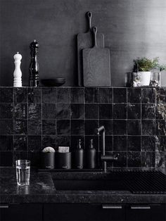 a kitchen sink with black tiles and cutting board on the wall above it, surrounded by other items