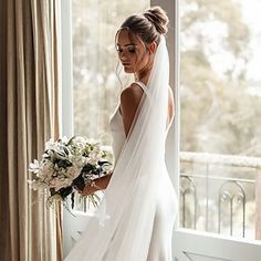 a woman standing in front of a window wearing a wedding dress and holding a bouquet