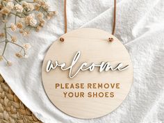 a wooden sign that says welcome please remove your shoes hanging on a white towel next to some flowers