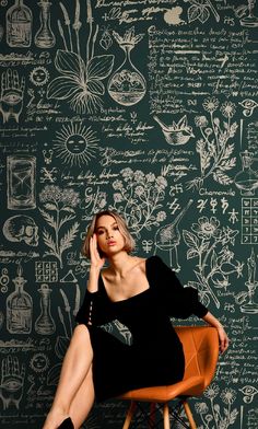 a woman sitting on top of a wooden chair in front of a wall covered with drawings