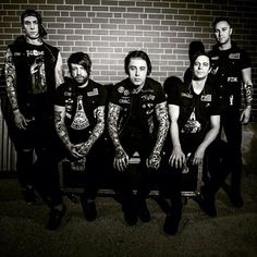 a group of men sitting next to each other in front of a brick wall with tattoos on their arms