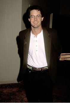 a man wearing glasses and a white shirt is standing in front of a door with his hands in his pockets