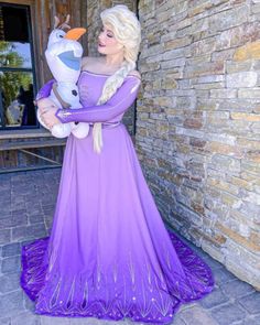 a woman in a purple dress is holding a frozen princess doll and posing for the camera