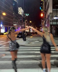 two women in sequin dresses crossing the street at night with their arms spread wide