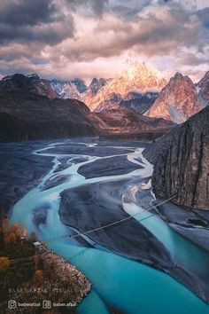 a river flowing through a valley surrounded by mountains under a cloudy sky with snow capped peaks in the distance