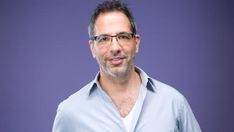 a man with glasses and a blue shirt is looking at the camera while standing in front of a purple background