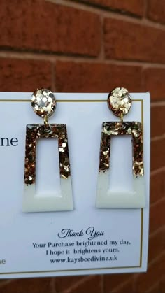 a pair of gold and white earrings on display in front of a brick wall with a card